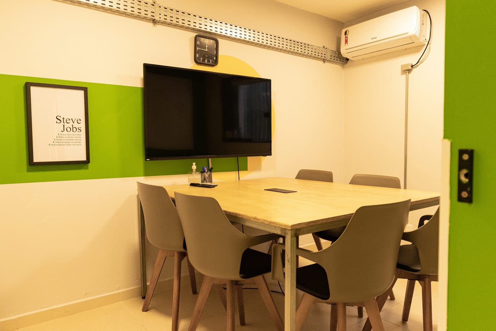 Sala de reunião do Brains Batel com parede verde e televisor, em Curitiba