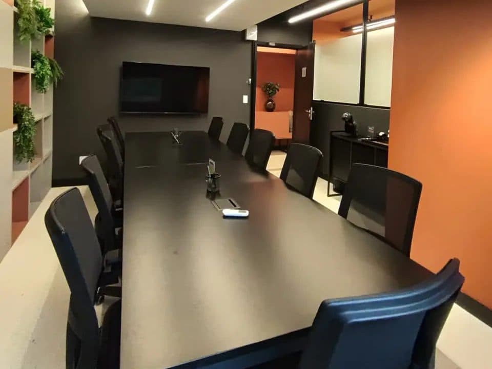 Sala de reunião do Brains Parque Barigui, com mesa longa para equipe, televisor e parede laranja, no Bigorrilho, em Curitiba
