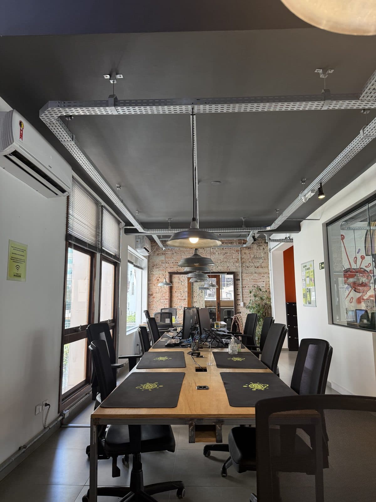Mesa comunitária do espaço compartilhado do Brains Batel, sob luminárias industriais, em Curitiba